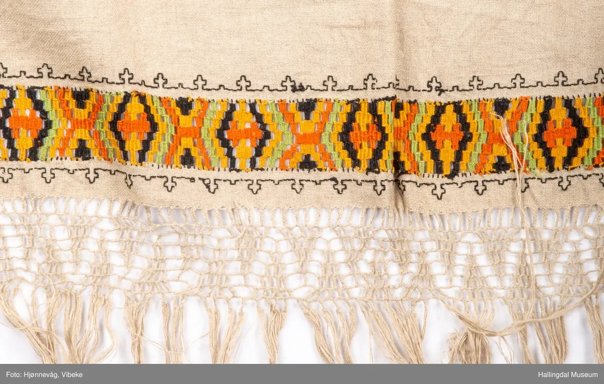 Vevd løper, toskaft i lin med frynser, beige. Broderi holbeinsøm. Nær frynsene er det broderi som ser ut som det er kelimvevd (uttrekksøm); svart, orange, sennep og grønt. Frynser med firflettet blonde i begge ender. Firflettingen er ikke gjort ferdig.