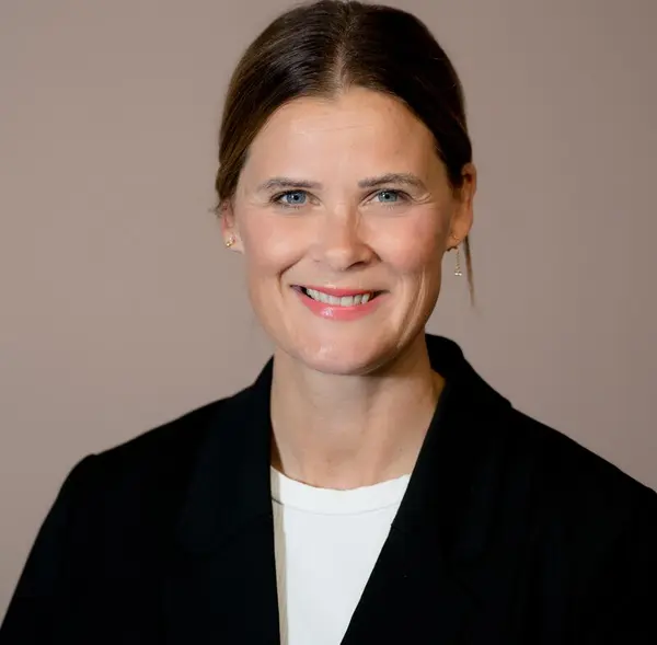 Portrettfoto av kvinne med mørkt hår. Håret er i midtskill og er satt opp. Hun har mørk dressjakke og hvit t-shirt. Hun smiler til kamera. Hun heter Cecilie Sørumshagen