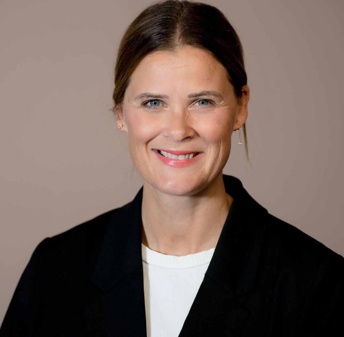 Portrettfoto av kvinne med mørkt hår. Håret er i midtskill og er satt opp. Hun har mørk dressjakke og hvit t-shirt. Hun smiler til kamera. Hun heter Cecilie Sørumshagen