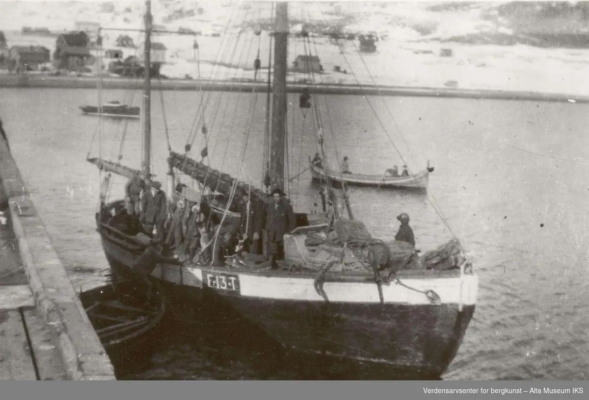 En seilbåt, F13T, ligger til kai i Talvik, og flere mennesker står i båten. Det er andre mindre båter bak dem i fjorden.