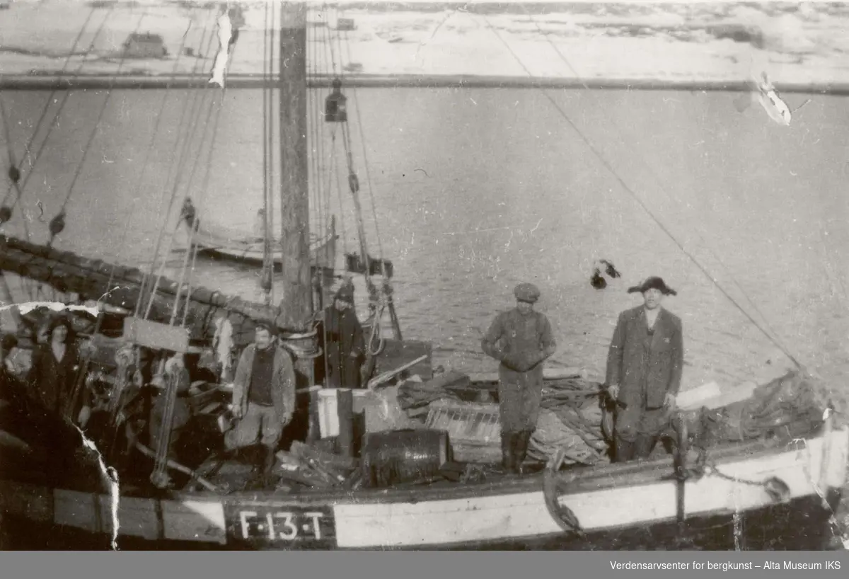 Flere mennesker står i en seilbåt, F13T,  som ligger til kai i Talvik. Bak båten ligger det andre mindre båter på fjorden.