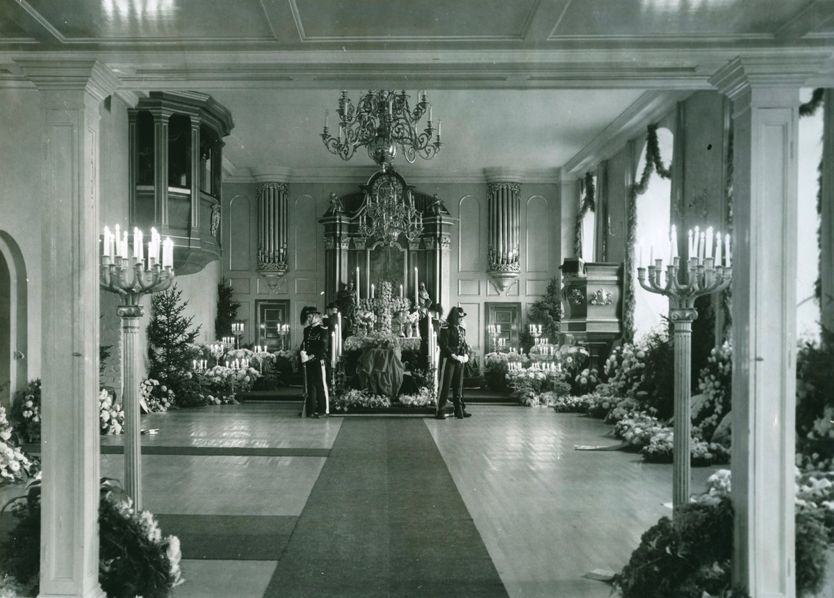 To gardister står æresvakt ved dronningens båre foran alteret i Akershus slottskirke. Blomster og tente lys. Kirkeinteriør.
