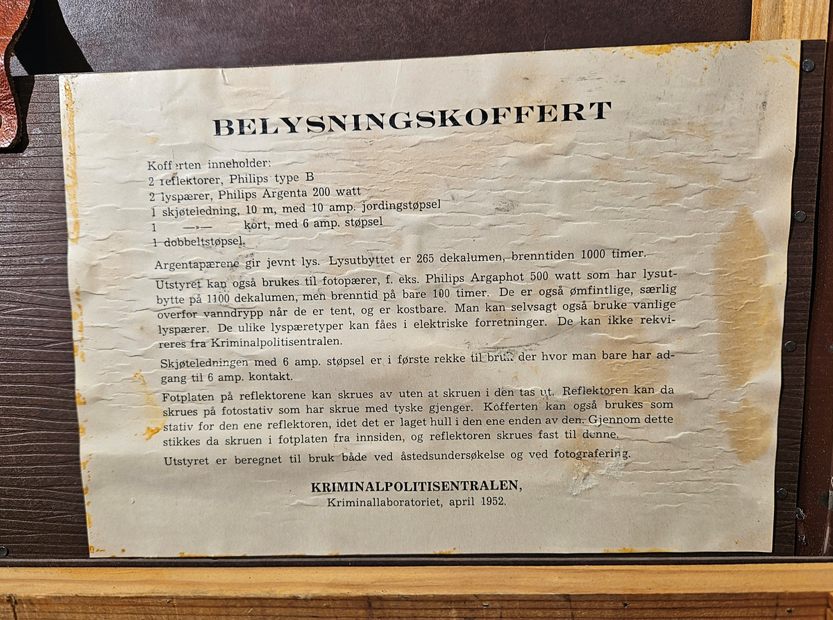 Brun koffert, forsterket i hjørnene. Kofferten inneholder en parafinlykt. Kofferten er brannskadet.