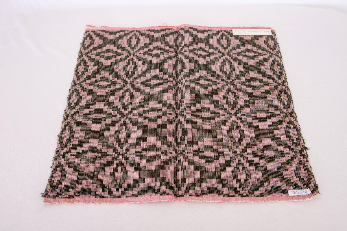 Möbeltygsprov vävt i jämtlandsdräll med oblekt bomullsgarn i varpen och grönt ullgarn i mönsterinslaget, rosa bomullsgarn  som mellaninslag.Provet har en påsydd bomullslapp med handskriven text: Bollnäs Hemslöjdsförening 79.En maskinskriven papperslapp med texten: Möbeltyg i jämtlandsdräll B.G 1920-talet är fästad vid provet.