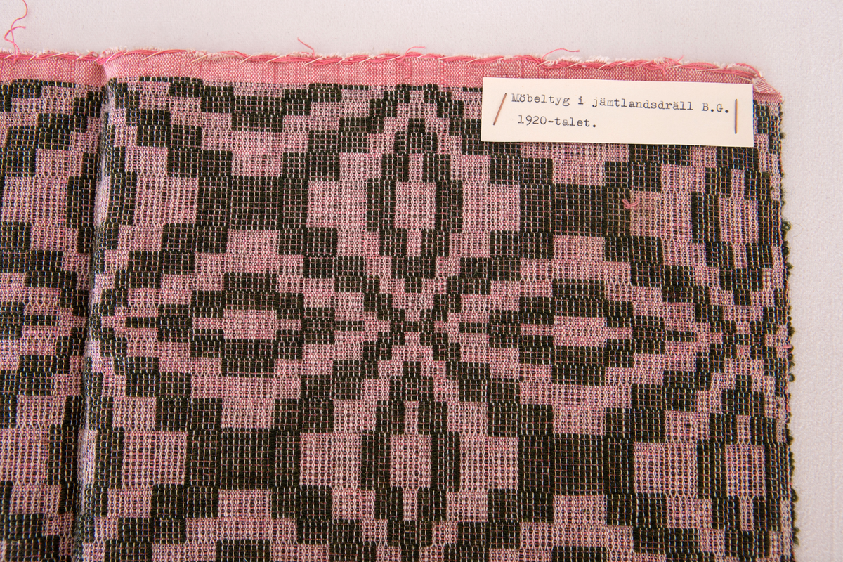 Möbeltygsprov vävt i jämtlandsdräll med oblekt bomullsgarn i varpen och grönt ullgarn i mönsterinslaget, rosa bomullsgarn  som mellaninslag.Provet har en påsydd bomullslapp med handskriven text: Bollnäs Hemslöjdsförening 79.En maskinskriven papperslapp med texten: Möbeltyg i jämtlandsdräll B.G 1920-talet är fästad vid provet.