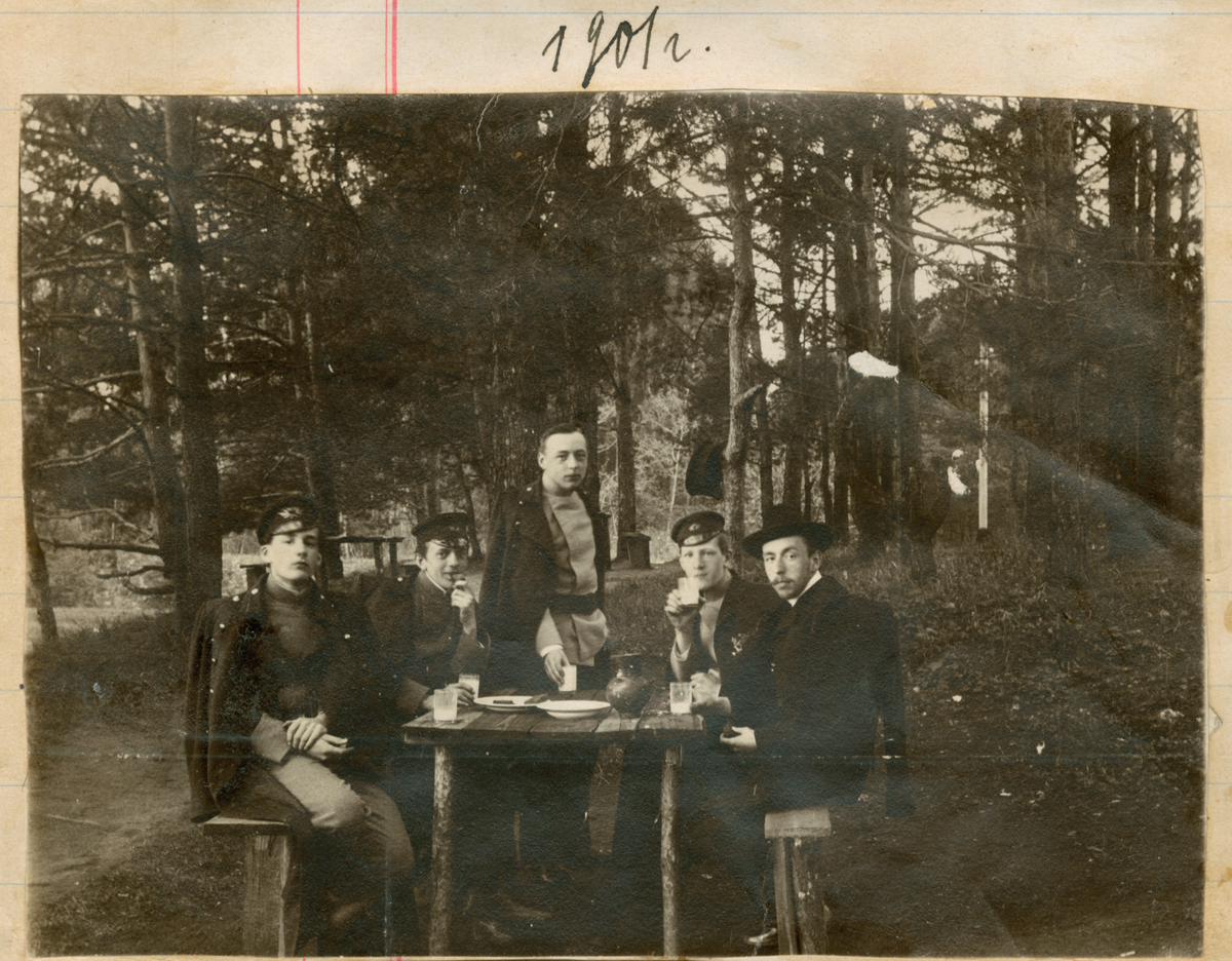 Fem menn fotografert under et måltid i skogen. De spiser og drikker melk på en benk. En hatt henger i treet.