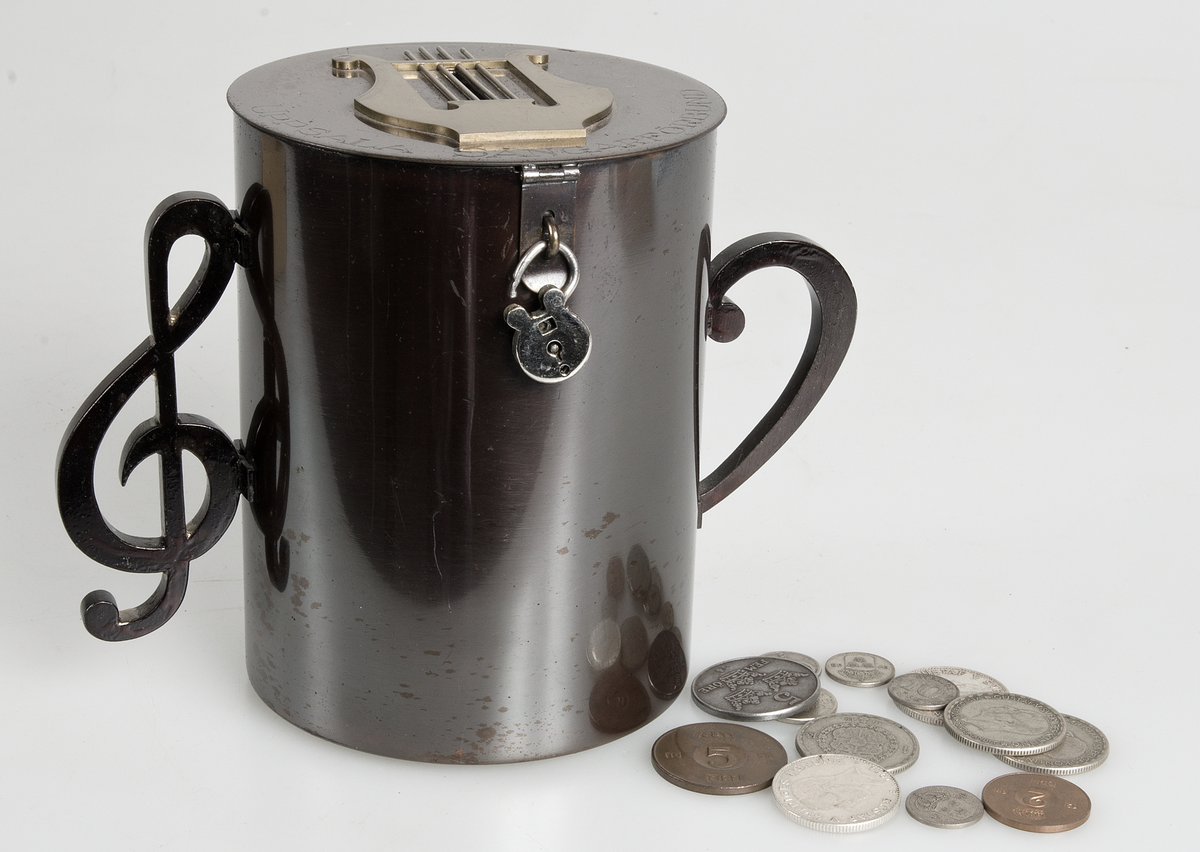 Sparbössa av brunerat stål (?), cylindrisk med platt lock på gångjärn, två handtag på sidorna i form av musikaliska tecken. På locket en lyra av gulmetall mellan vars strängar myntinkastet är samt graverad text "UPSALA SÅNGARFÖRBUND". Litet hänglås. På lockets insida signerad H.M. 1928.