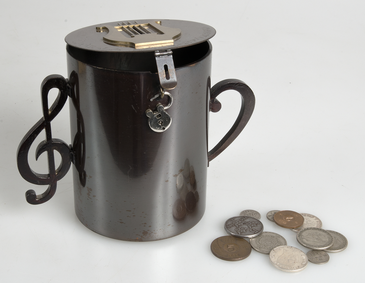 Sparbössa av brunerat stål (?), cylindrisk med platt lock på gångjärn, två handtag på sidorna i form av musikaliska tecken. På locket en lyra av gulmetall mellan vars strängar myntinkastet är samt graverad text "UPSALA SÅNGARFÖRBUND". Litet hänglås. På lockets insida signerad H.M. 1928.
