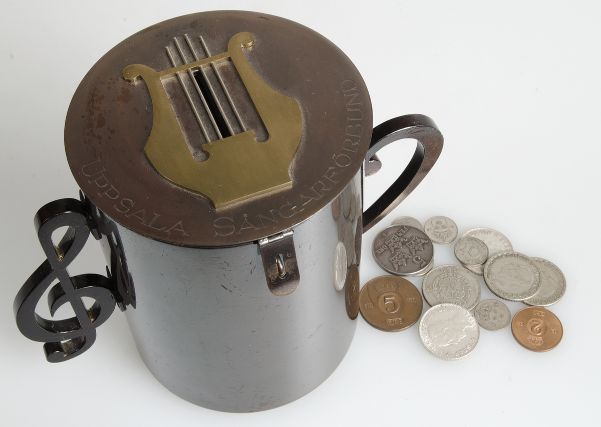 Sparbössa av brunerat stål (?), cylindrisk med platt lock på gångjärn, två handtag på sidorna i form av musikaliska tecken. På locket en lyra av gulmetall mellan vars strängar myntinkastet är samt graverad text "UPSALA SÅNGARFÖRBUND". Litet hänglås. På lockets insida signerad H.M. 1928.