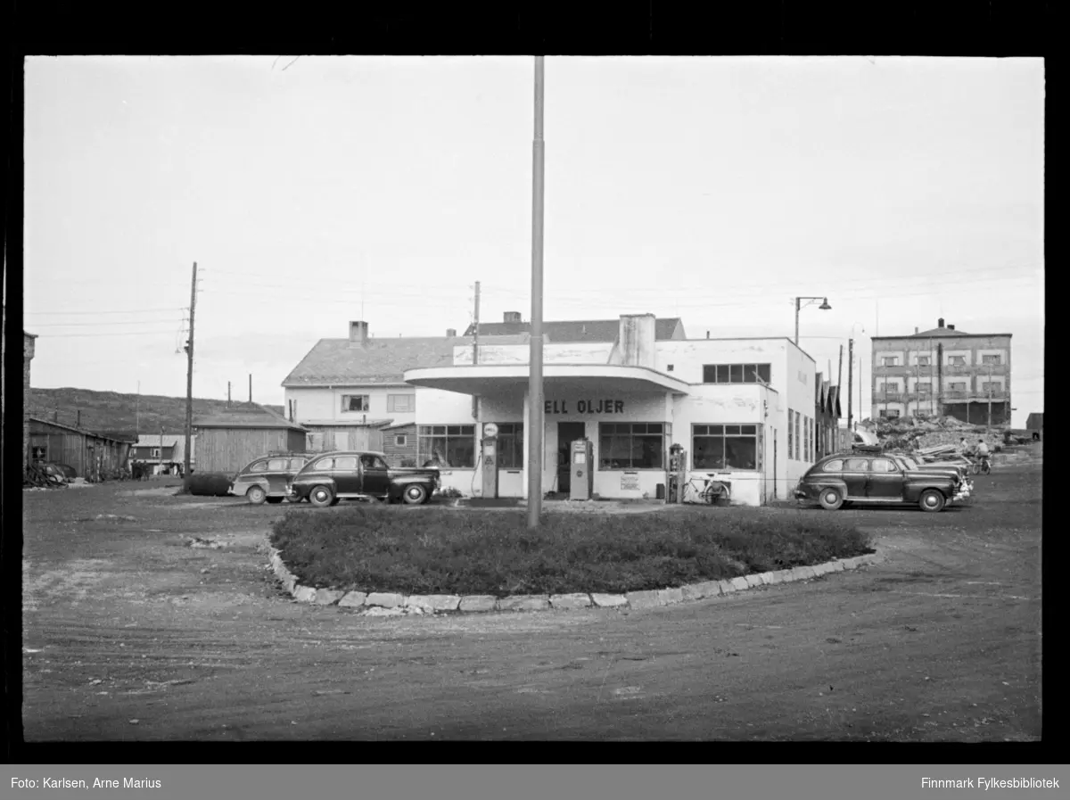 Foto av Shell Oljer bensinstasjon i Kirkenes 

I foto kan man se at gaten er en grusvei. Ved siden av bensinstasjonen ser man flere biler parkert og foran stasjonen er bensinpumper. 