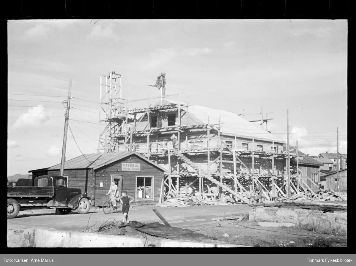 Bygg konstrueres i Kirkenes etter krigen. Ved siden av står et mindre hus med skilt "O. M. Hatle & Co."