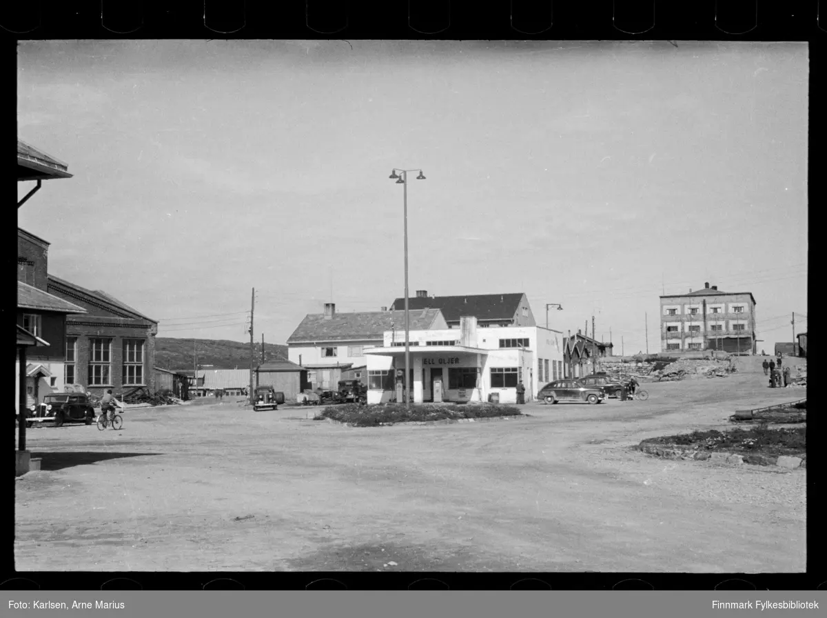 Foto av Shell Oljer bensinstasjon i Kirkenes 

I foto kan man se at gaten er en grusvei. Ved siden av bensinstasjonen ser man flere biler parkert og foran stasjonen er bensinpumper. 