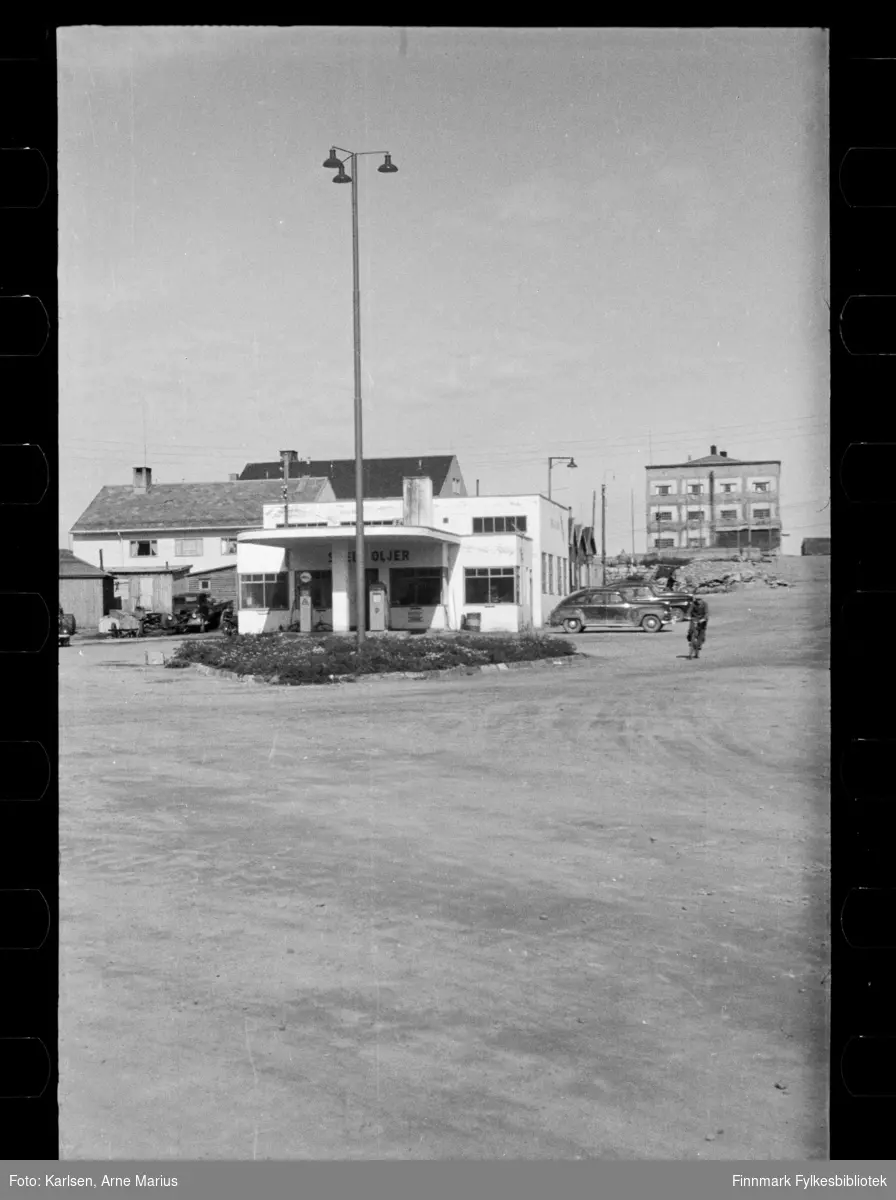 Foto av Shell Oljer bensinstasjon i Kirkenes 

I foto kan man se at gaten er en grusvei. Ved siden av bensinstasjonen ser man flere biler parkert og foran stasjonen er bensinpumper. 