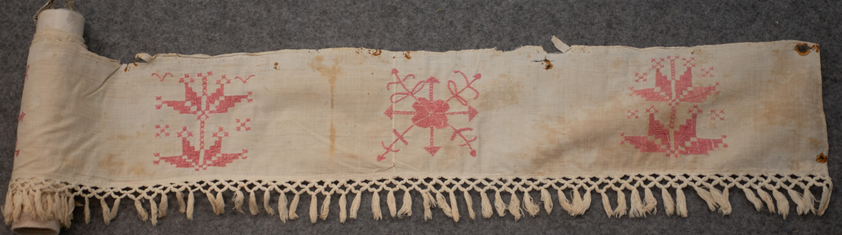 Hängkläde av bomullstyg med broderier av bomullsgarn. Vit botten och rödabroderier. Märkt "C S E D". Längd 243 cm. Bredd 16 cm. Bredd (Frans) 6 cm. Frans 50 mm.