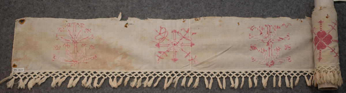 Hängkläde av bomullstyg med broderier av bomullsgarn. Vit botten och rödabroderier. Märkt "C S E D". Längd 243 cm. Bredd 16 cm. Bredd (Frans) 6 cm. Frans 50 mm.