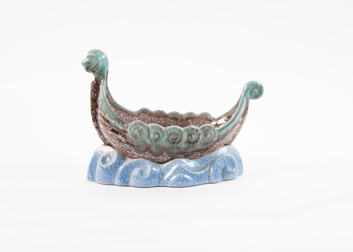 Lampefot formet som vikingskip. Stett formet som bølger. Tydelig skjoldrekke på sidene, bakovervendt hale. Hvit bunnglasur med blå overglasur på stett.