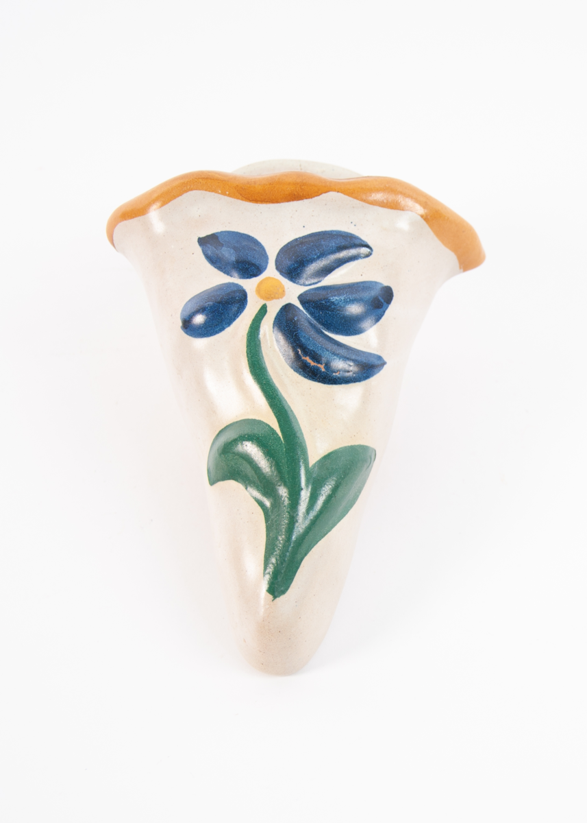 Kremmerhusformet vegghengt blomstervase. Beige bunnglasur med lys brunoransje kant. Blomster relieff i front i grønt, blått og gult.