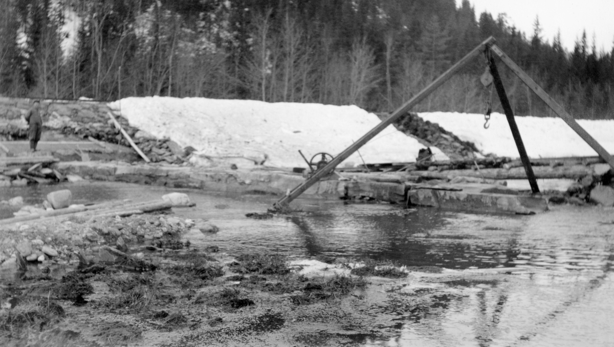 Bygging av strømterskel i elva Imsa i Stor-Elvdal i Hedmark i 1939. Fotografiet er tatt mot en mur hvor det sto en stubbebryter, et trebeint stativ med talje under krysniingspunktet i toppen. Dette redskapet ble brukt til å løfte tunge steinblokker opp på muren som var under oppførelse. Helt til venstre i bildet, ved flomvollen langs elveløpet, sto det en arbeidskledd mann.