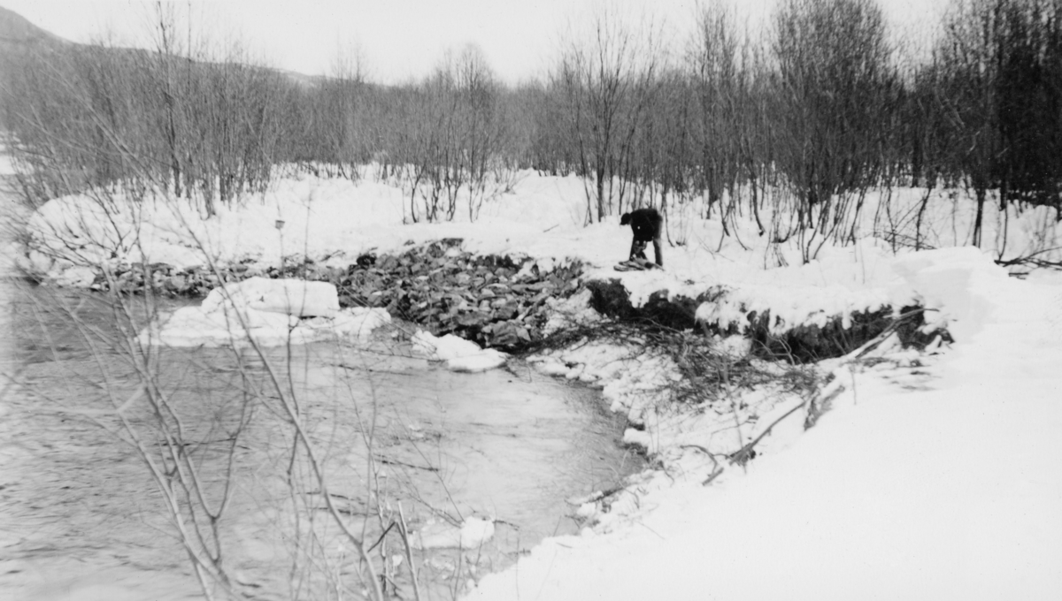 Forsterkning av en av yttersvingene i elva Nordre Eldåa i Stor-Elvdal vinteren 1939. Vi ser en mann som bærer forholdsvis grov stein til elveskråningen for å hindre at vannet i elva skal grave vekk løsmasser og på den måten gjøre svingen stadig krappere, noe som ville skapt problemer under tømmerfløtinga. Arbeidet ble utført mens det lå snø på marka og mens vannføringa i Nordre Eldåa var forholdsvis lav. 

Gardsnavnet Akselstu er av en eller annen grunn knyttet til dette fotografiet, Kanskje bildet er tatt på et utmarksareal som tilhørte denne eiendommen?