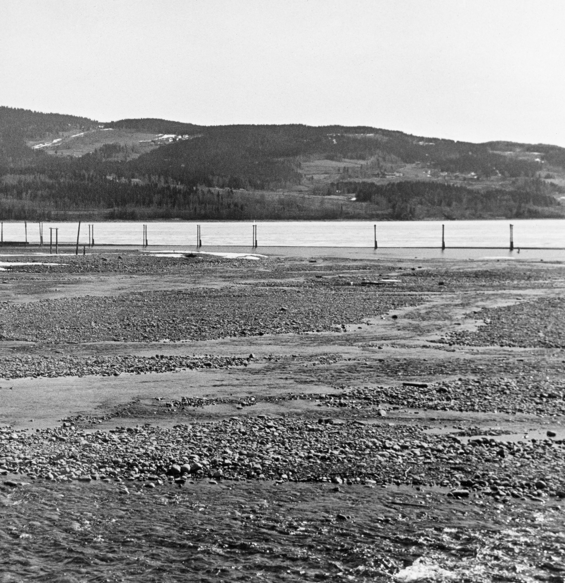 Fra elva Rindas utløp i Mjøsa ved Vingrom i daværende Fåberg kommune i Oppland.  Fotografiet er tatt våren 1939.  Vi ser ut over et bredt, grunt og steinete elveløp med lav vannføring og enkelte små isflak etter foregående vinter.  På motsatt side av Mjøsa ser vi i retning bygdelaget Søre Ål, som også lå i Fåberg. I Glomma fellesfløtingsforenings album er dette bildet er montert som den venstre delen av et todelt panoramaopptak.  

Elva Rinda kommer fra Rindtjernene sør for Rindåsen, i grensetraktene mellom Vestre Gausdal og Fåberg.  Den renner gjennom Saksumdalen, og når Mjøsa etter drøyt to mil.  Rinda renner gjennom skogtrakter, og den var derfor ei viktig fløtingselv.  For å hindre at fløtingsvirket strøk fra elveosen og ut i Mjøsa ble det oppsatt ei lense som skulle samle opp alle stokkene, slik at de kunne buntes før de ble buksert videre sørover.  Lensa besto av en kjede av flytende tømmerstokker, støttet av ei pælerekke.
