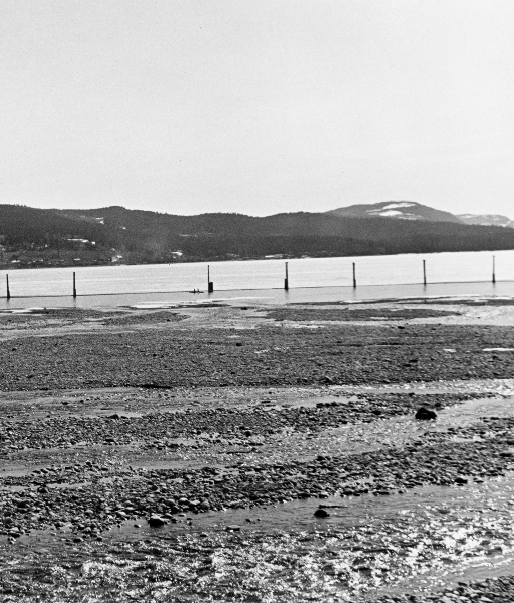 Fra elva Rindas utløp i Mjøsa ved Vingrom i daværende Fåberg kommune i Oppland.  Fotografiet er tatt våren 1939.  Vi ser ut over et bredt, grunt og steinete elveløp med lav vannføring og enkelte små isflak etter foregående vinter.  På motsatt side av Mjøsa ser vi i retning bygdelaget Søre Ål, som også lå i Fåberg. I Glomma fellesfløtingsforenings album er dette bildet er montert som den høyre delen av et todelt panoramaopptak.  

Elva Rinda kommer fra Rindtjernene sør for Rindåsen, i grensetraktene mellom Vestre Gausdal og Fåberg.  Den renner gjennom Saksumdalen, og når Mjøsa etter drøyt to mil.  Rinda renner gjennom skogtrakter, og den var derfor ei viktig fløtingselv.  For å hindre at fløtingsvirket strøk fra elveosen og ut i Mjøsa ble det oppsatt ei lense som skulle samle opp alle stokkene, slik at de kunne buntes før de ble buksert videre sørover.  Lensa besto av en kjede av flytende tømmerstokker, støttet av ei pælerekke.