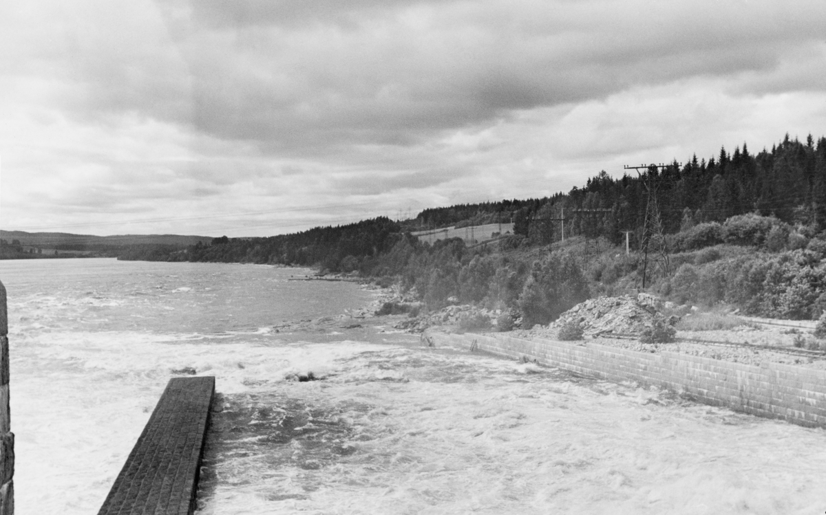 Fra damanlegget ved Rånåsfoss i Sørum kommune på Romerike. Dette fotografiet viser ei bred, plan, men nedoverskrånende betongrenne, antakelig under ei av sektorlukene på den 186 meter lange dammen. Sjøl om dette kraftverket hadde ei tømmerrenne på vestre elvebredd som skulle kunne ta unna om lag 180 000 tømmerstokker per døgn med ei vannføring på 15 til 30 kubikkmeter i sekundet, ble tømmeret vanligvis lenset i retning av de nevnte sektorlukene. Dette skyldtes ei bakevje ved renneinntaket, noe som gjorde påstikkinga svært mannskapskrevende. Den egentlige tømmerrenna (se SJF.1994-01064) ble rett og rett og slett revet i 1962 (se SJF.1990-02174). Dette fotografiet ble tatt i 1939.