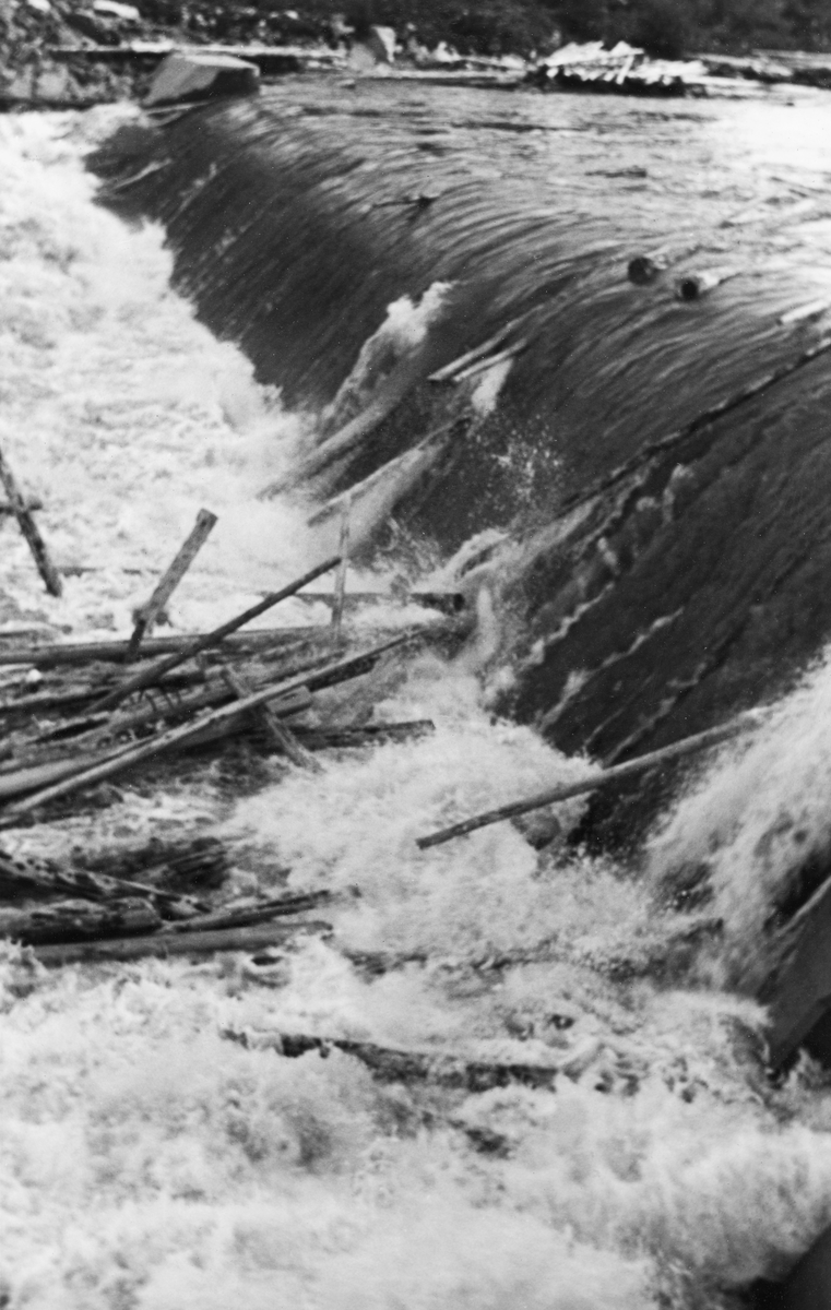 Fra dammen ved Kvernfallet i elva Søndre Osa, like ved gardsbruket Ulvåvollen i Åmot.  Fotografiet ble tatt under fløtinga i 1939, fra sørsida av vassdraget, skrått mot dammen. Kraftverket som hentet sin energi fra dette fallet ble satt i drift i 1936. Dammen som skulle stuve opp vann til den nedenforliggende kraftstasjonen ser ut til å ha skapt en bortimot to meter høy, avrundet terskel i vassdraget.  Da bildet ble tatt var elva preget av vårflom om mye fløtingsvirke.  Vi ser hvordan stokkene gled over damkrona mot den kvitskummende sonen like nedenfor, der det ser ut til at det var i ferd med å bygge seg opp en tømmervase.