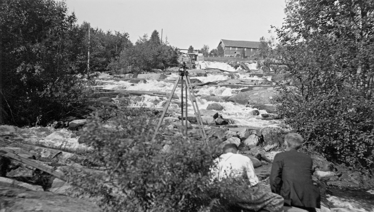 Dette fotografiet skal være tatt i Kråkfossen eller Krokfossen i elva Leira, som renner gjennom Nannestad på Romeriket. Vi ser fossen i motstrøms retning. I forgrunnen på dette bildet står det et trebeint stativ, antakelig med en nivelleringskikkert. Høyere oppe fossen ser vi ei vegbru og på bakkekammen ligger driftsbygningen på en gard.