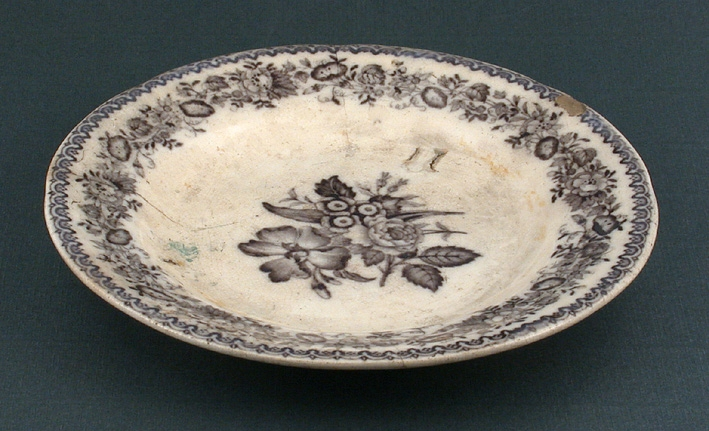 Vit assiett med rik blomdekor i mörkt brun färg runt kanten och i assiettens mitt. Fabrikat och märkning oklart då stämplar är oläsliga.