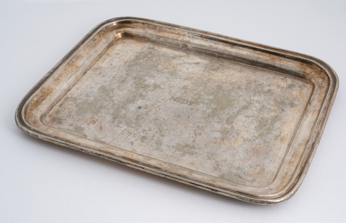 To identiske serveringsfat av metall fra NFDS (Nordenfjeldske Dampskibsselskap). De er produsert av Walker & Hall. Fatene er firkantede med avrundede hjørner. Serveringsdelen har samme form og måler 33,5x25,5cm. Midt på fatene er "N.F.D.S." gravert inn/stemplet inn. På undersiden er det innskrift med informasjon om produksjon, se "Påført tekst/merker".