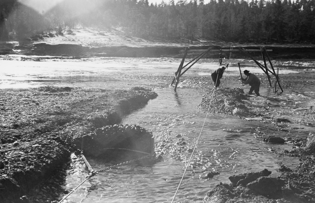 «Elveforbedringsarbeid» med slepeskrape som ble manøvrert ved hjelp av en bakmontert vinsj på en beltetraktor - traktoren med vinsjen er avbildet på SJF.1989-03528. Her har fotografen vendt kameraet den andre vegen, mot ei stålskuffe, som samtidig med at de trekkes mot traktoren med vinsjene fylles med elvegrus, som på denne måten trekkes på land. Vaierne fra vinsjen gikk via ei talje som var motert i en høy bukk som var oppstilt vinkelrett på trekkretningen,  Like framfor denne bukken arbeidet to karer med hakker, antakelig for å forberede neste drag med skrapeslkuffen.