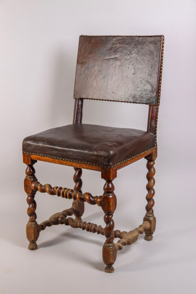 Stol tillverkad av björk, i barockstil. Ryggbricka, ryggstolpar och sits klädda i skinn, fäst med nitar. Fyra raka ben, med stabiliserande framslå, samt ett stabiliserande H-format benkryss. Ben och slår delvis profilsvarvade och delvis spiralsvarvade. Benen med konformade fotavslut. 