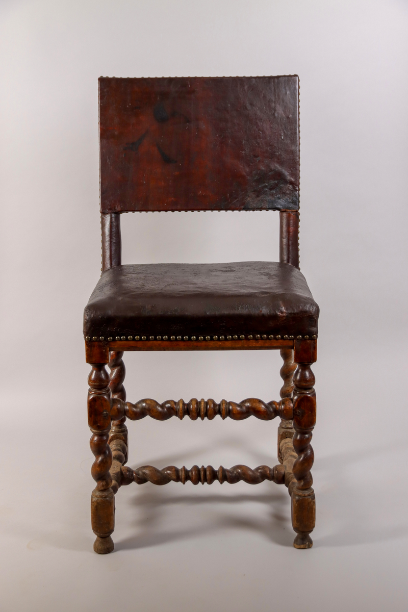 Stol tillverkad av björk, i barockstil. Ryggbricka, ryggstolpar och sits klädda i skinn, fäst med nitar. Fyra raka ben, med stabiliserande framslå, samt ett stabiliserande H-format benkryss. Ben och slår delvis profilsvarvade och delvis spiralsvarvade. Benen med konformade fotavslut. 