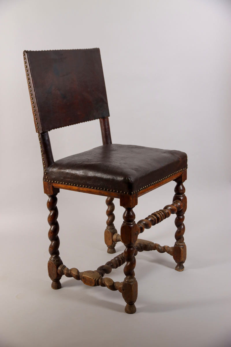 Stol tillverkad av björk, i barockstil. Ryggbricka, ryggstolpar och sits klädda i skinn, fäst med nitar. Fyra raka ben, med stabiliserande framslå, samt ett stabiliserande H-format benkryss. Ben och slår delvis profilsvarvade och delvis spiralsvarvade. Benen med konformade fotavslut. 