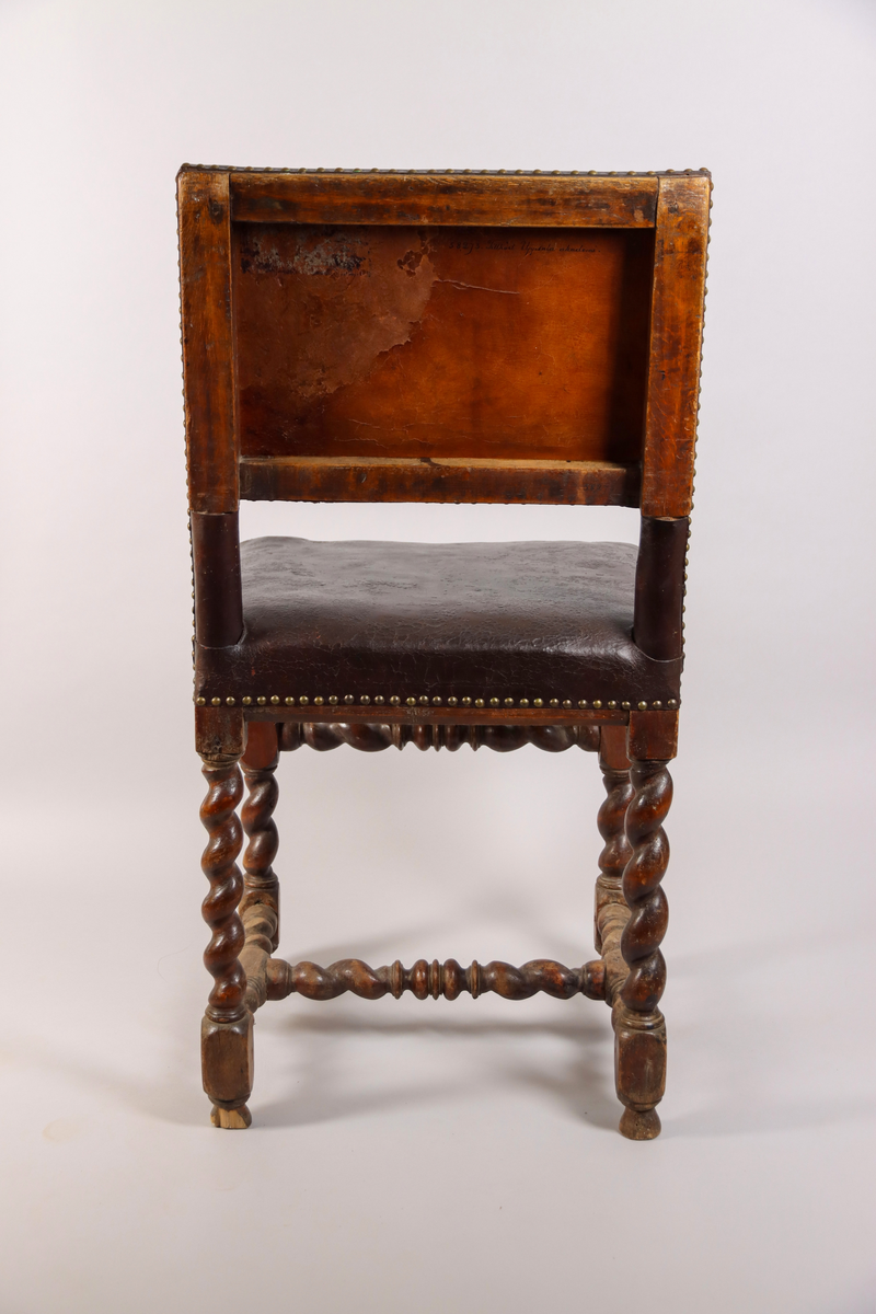 Stol tillverkad av björk, i barockstil. Ryggbricka, ryggstolpar och sits klädda i skinn, fäst med nitar. Fyra raka ben, med stabiliserande framslå, samt ett stabiliserande H-format benkryss. Ben och slår delvis profilsvarvade och delvis spiralsvarvade. Benen med konformade fotavslut. 