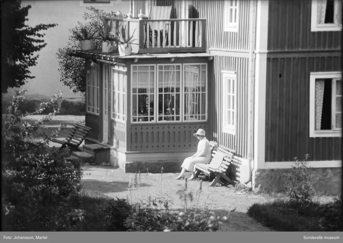 Två bilder av en bostadsbyggnad med glasveranda. På en bänk sitter en kvinna i ljus dräkt och hatt. Den ljusa byggnaden bakom är samma som SuM-foto034836, Villa Göta. Okänt var i Sverige bilden är tagen.