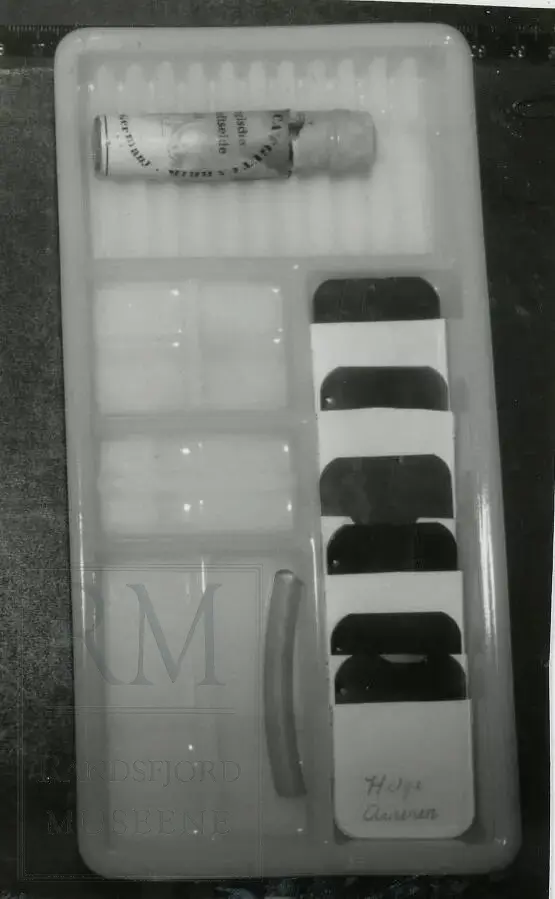 Skålen er delt i 5 store og mindre rom. Inneholder 6 røntgenbilder, steril tråd, 1 flaske med skrukork og 1 bit plastslange. Røntgenbildene ligger i små hvite plastmapper med påskrevne navn.