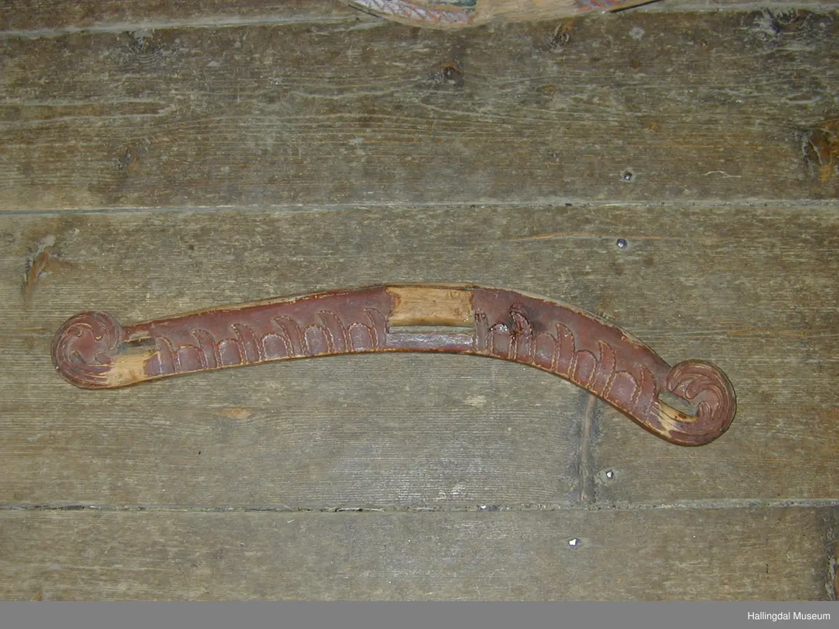 Eit krumt trestykke med eit rektangulært hol i kvar ende og eit på midten. Skore ornamentikk på eine sida (bladornamentikk), skore ei slags "opprulling" ytst i kvar ende.
