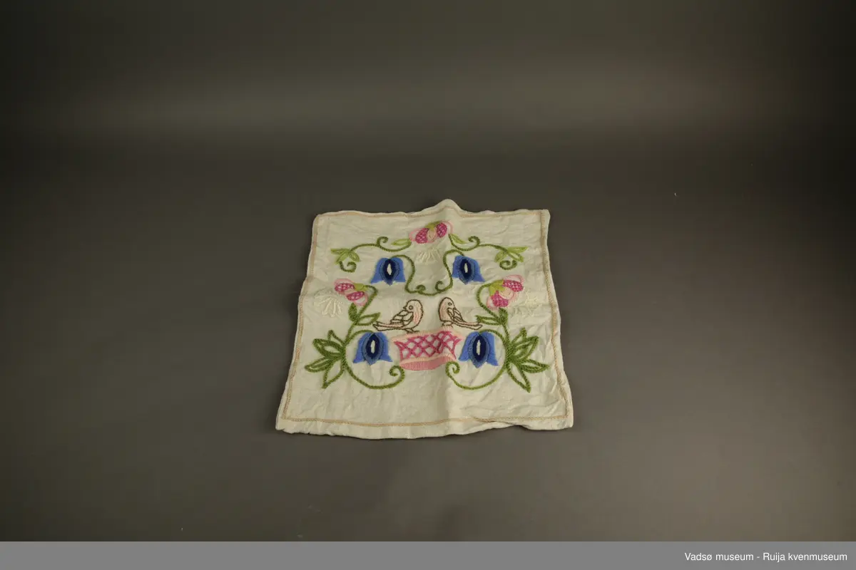Beige putetrekk med et vakkert broderimotiv på fremsiden. Motivet viser to fugler som er omgitt av blå og rosa blomster. Rundt motivet er det brodert en beige kant.