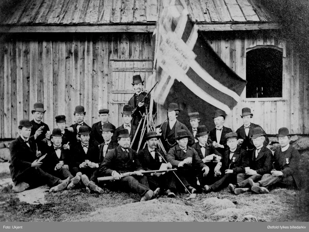 Rakkestad skytterlag og medlemmer av Eidsberg skytterlag  foran skytterhuset på Søndre Fladstad i Rakkestad i 1880 (1880-åra). I lagets historie er det oppgitt at bildet er fra skytterhuset på Schiesmyren. Det er Rakkestad Skytterlags flagg (med sildesalat) som vaier i bakgrunnen. Skytterlaget ble stifta 17. mai i 1861 med navnet Rakkestads Skydeselskab. Skytterhus ble oppført i 1863, og bane anlagt. 
På bildet er det også skyttere fra Eidsberg skytterlag som gjester skytterlag i Rakkestad.. Karene koser seg med en dram og er pynta med medaljer, det kan være et stevnearrangement.
Eidsberg skytterlag blei stifta 17. mai 1858 på Mustorp. 

Navneopplysning fra nesten identisk motiv publisert på Oslo Museums side: "Bak, nr. 3 fra høyre er verkseier Hans Martin Torp. Foran nr. 3 fra høyre er K. E. Jahren. (Kristian Edvard Jahren)". Begge fra Rakkestad.

Bygdebok for Eidsberg 1940. s. 451, har oppgitt disse navnene for samme bilde,  altså ant navn på de som var fra Eidsberg (?): 
"Mannen lengst til venstre er Ole Torkildsen Filtvedt. De to i midten foran - med geværet over knærne - er en folkehøyskolelærer (enten Peder Velde (f. 1860) eller Lasse Trædal f. 1857 - må være P. Velde) og Ingebrigt (?) Præsthus (med medalje på brystet). Lengst til høyre sitter Johannes Spestad. Bak til venstre sitter Nils Granberg, og nummer to fra høyre i bakerste rekke er Anders Torp fra Trømborg."