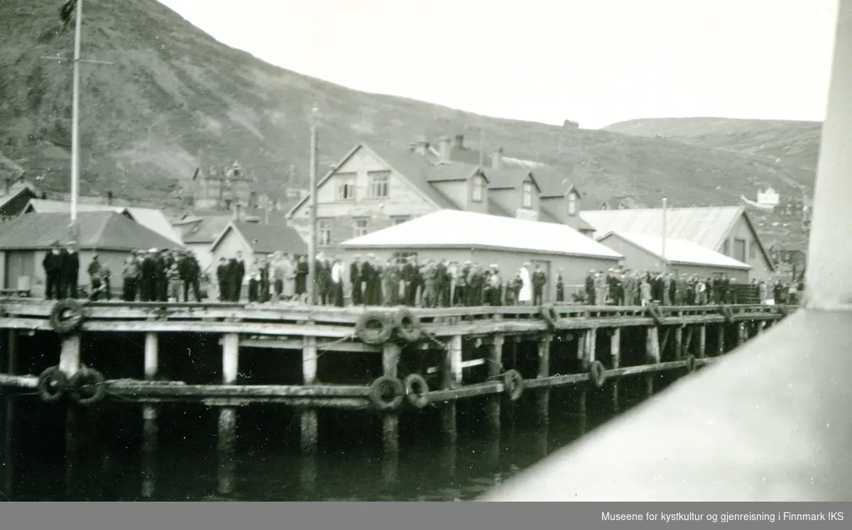 Honningsvåg. Dampskipskaia med bebyggelse og mange personer. Bilde er tatt fra et skip. Antatt 1930-årene.