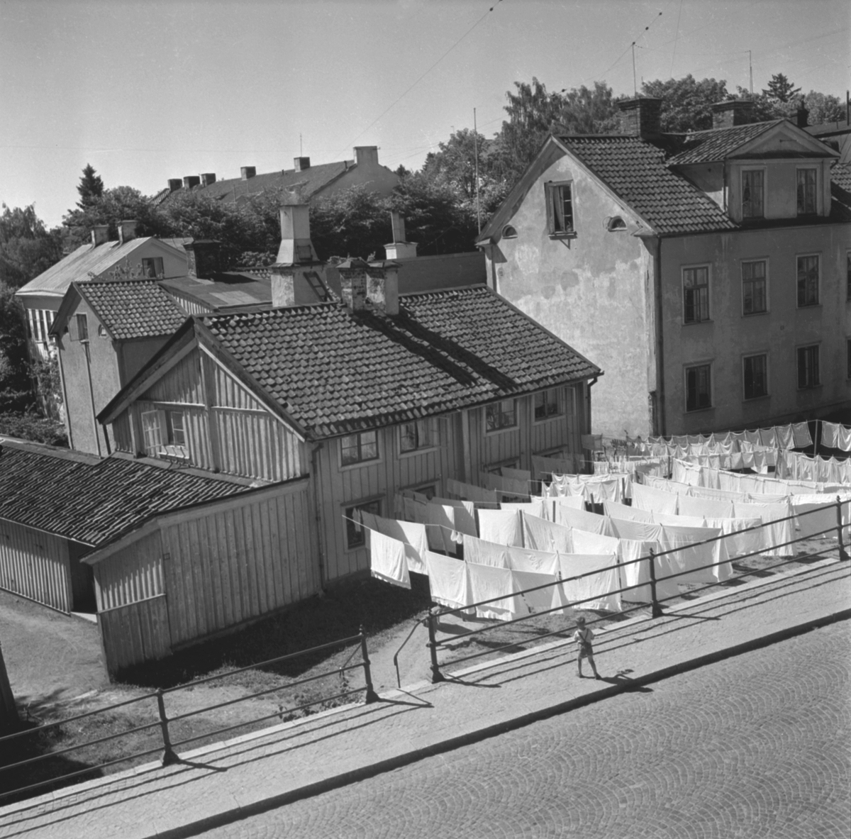 Utsikt mot Nygatan 62, Anderska gården, i Linköping omkring 1950. Anderska gården, likt övriga utmed stråkets södra sida, låg betydligt lägre än gatunivån. Läget förklaras av att husen i ett tidigare skede legat utmed en äldre sträckning av Sankt Korsgatan, som under 1870-talet lagts igen när Nygatan förlängdes västerut. Just denna dag rådde för övrigt torkväder, vilket gladde tvätterskan Selma Johansson som fyllt gårdsplanen med vittvätt.