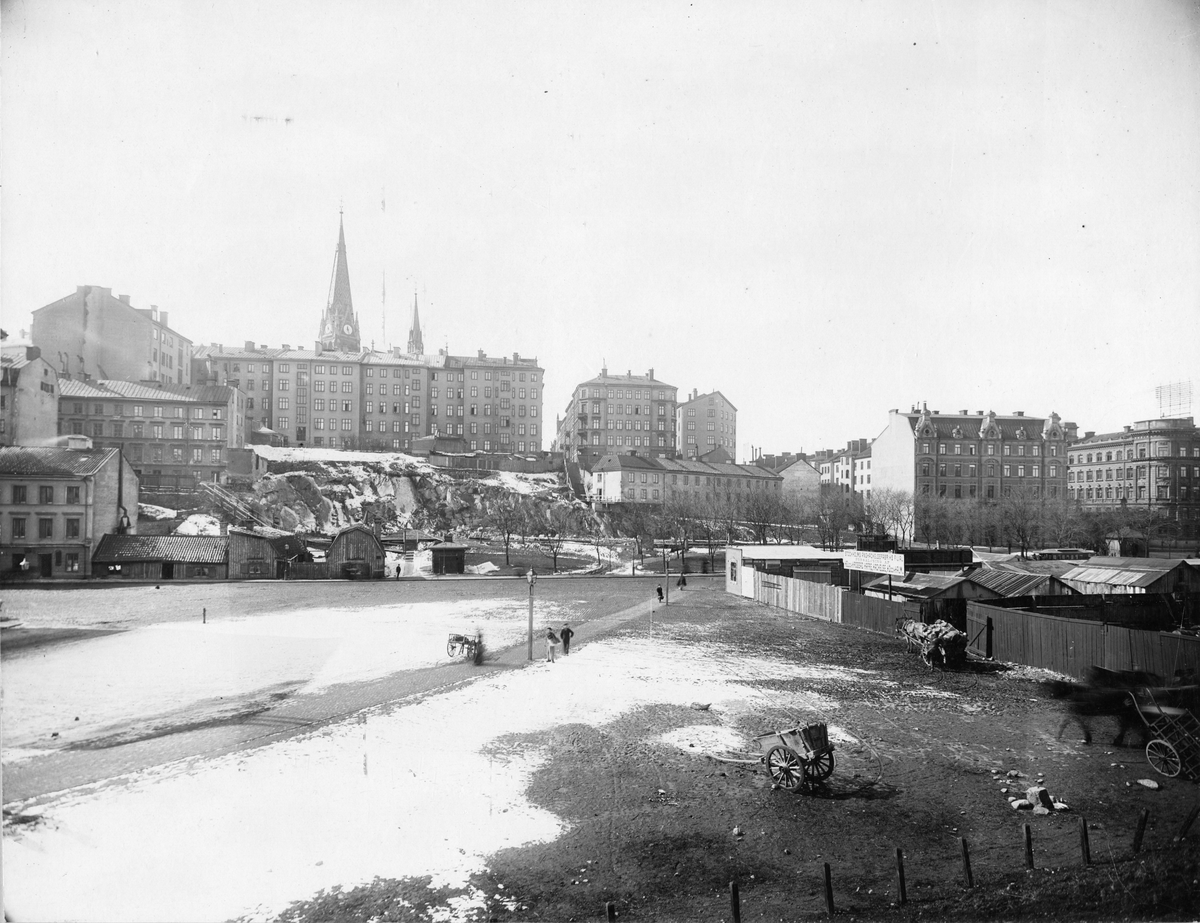 Vy från Stockholm. Stockholms Ånghackelsefabrik. I bakgrunden syns Sankt Johannes kyrka.
Text på skylt: "Stockholms Ånghackelsefabriks filial. Försäljning af helpressad hafre, hackelse, hö och halm."