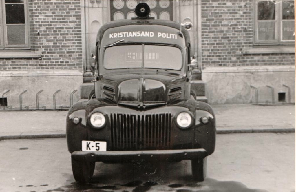 Cellebil, Ford 1946-modell, med registreringsnummer K-5 parkert foran en bygning. Bilen er merket "Politi Kristiansand S.", og er fotografert både forfra og fra siden.