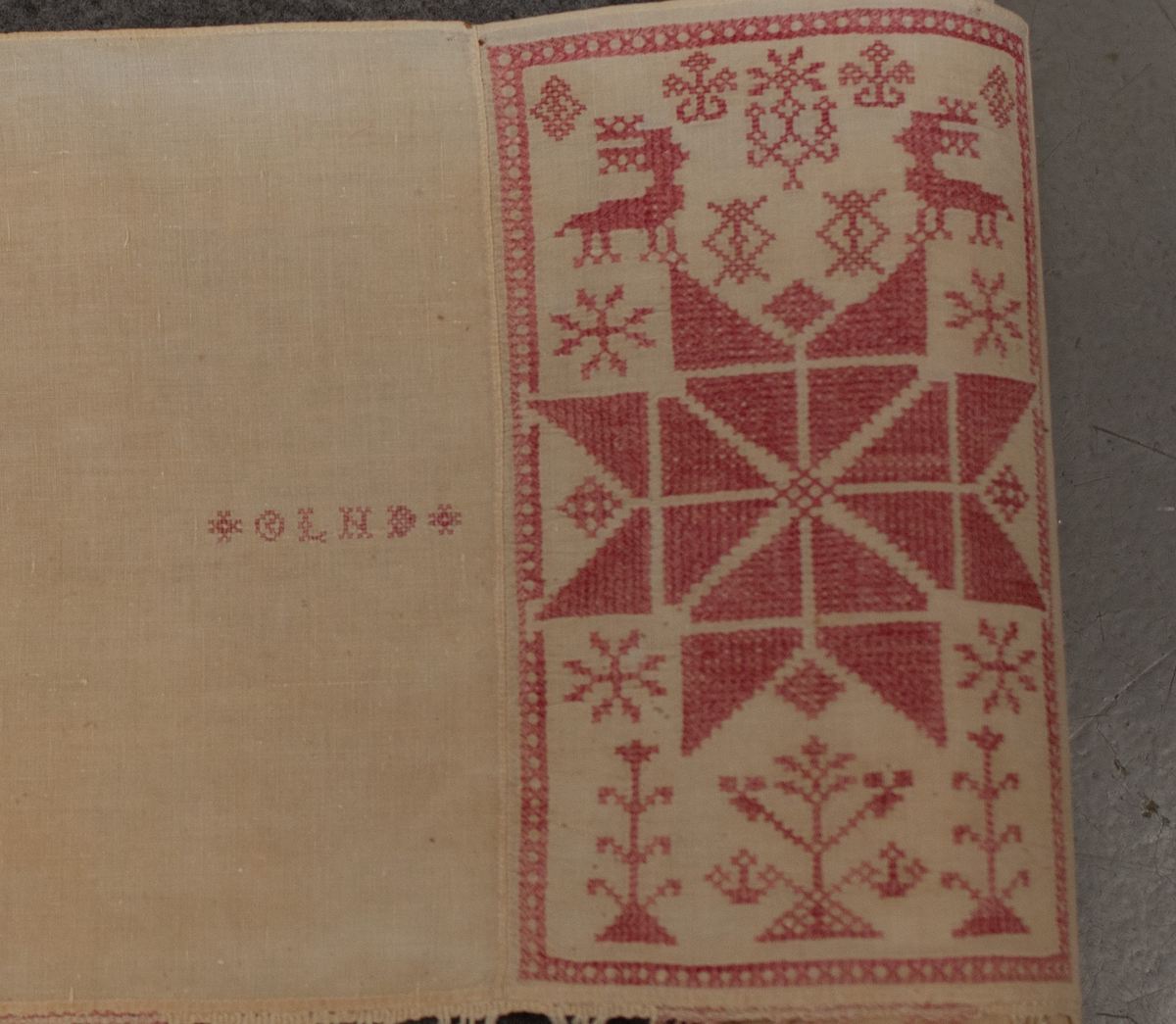 Hängkläde av halvlinne med bomullsgarn till korsstyngnbroderierna. Vit botten, broderier i rött. Knuten frans i rött. Längd 589 cm. Bredd 27 cm. Frans 8 cm. Märkt "1850", "O L N D". Frans: 70 mm.