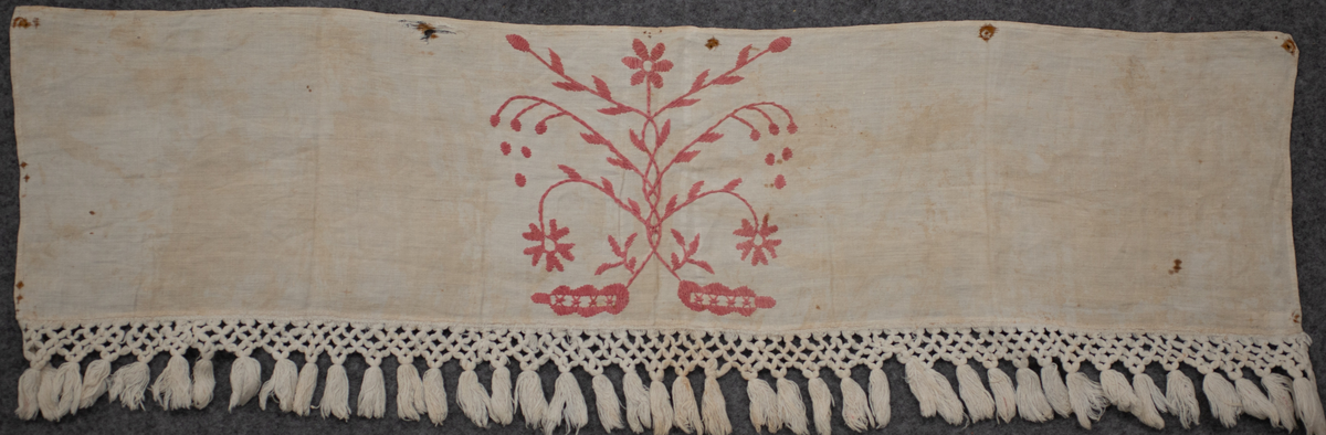 Hängkläde, del av. En liten bit av ett broderat hängkläde med hallandssöm. Bottentyg i tuskaftat vitt linne med ljusrött (rosa) broderi. Påsydd vit frans av bomull med omknutna tofsar i nederkanten. Längd= 100 cm. Bredd= 23,5 cm. Längd (frans)= 8,5 cm.