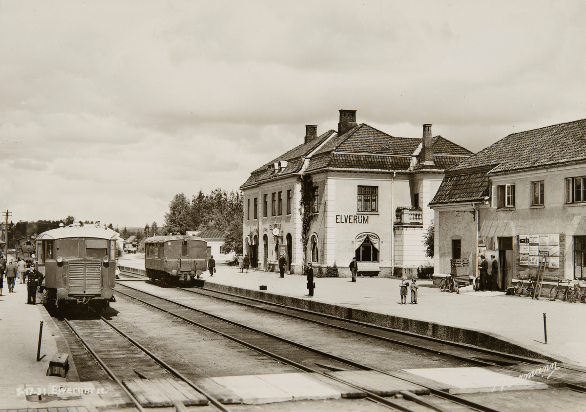Postkort, Elverum stasjon, jernbanestasjon, stasjonsbygning, jernbanespor, lokomotiv, reisende på perrongen, godshus med reklameskilter,