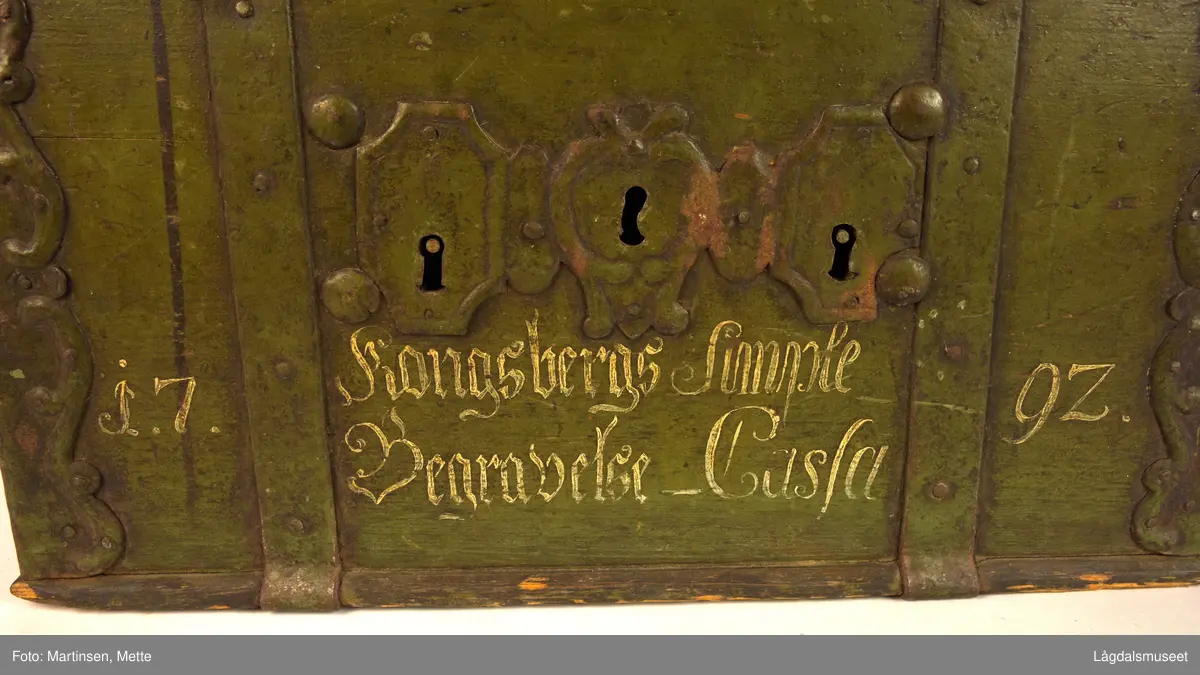 Solid, jernbeslått kiste med buet lokk. Hjørnebeslag av jern med C-ornamenter. På hver kortside jernhåndtak. Tre nøkkelhull ved siden av hverandre på forsiden med beslag. Beslagene er satt på med nagler og solide, grove bolter. På forsiden påmalt med hvitt, årstallet 1792 og "Kongsbergs Simple Begravelse-Casse". Kista er grønnmalt (olivengrønn), avslitt på lokket.