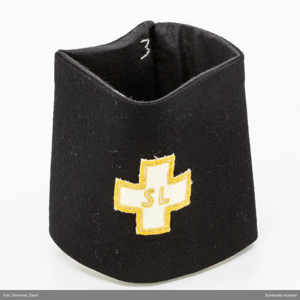 En svart armbindel som bär ett vitt kors med guldkolorerade bokstäver samt kantdekoration.