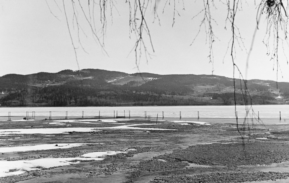 Fra elva Rindas utløp i Mjøsa ved Vingrom i daværende Fåberg kommune i Oppland.  Fotografiet er tatt våren 1939.  Vi ser ut over et bredt, grunt og steinete elveløp med lav vannføring og enkelte små isflak etter foregående vinter.  På motsatt side av Mjøsa ser vi i retning bygdelaget Søre Ål, som også lå i Fåberg. I Glomma fellesfløtingsforenings album er dette bildet montert som høyre del av et todelt panoramaopptak. 

Elva Rinda kommer fra Rindtjernene sør for Rindåsen, i grensetraktene mellom Vestre Gausdal og Fåberg.  Den renner gjennom Saksumdalen, og når Mjøsa etter drøyt to mil.  Rinda renner gjennom skogtrakter, og den var derfor ei viktig fløtingselv.  For å hindre at fløtingsvirket strøk fra elveosen og ut i Mjøsa ble det oppsatt ei lense som skulle samle opp alle stokkene, slik at de kunne buntes før de ble buksert videre sørover.  Lensa besto av en kjede av flytende tømmerstokker, støttet av ei pælerekke.